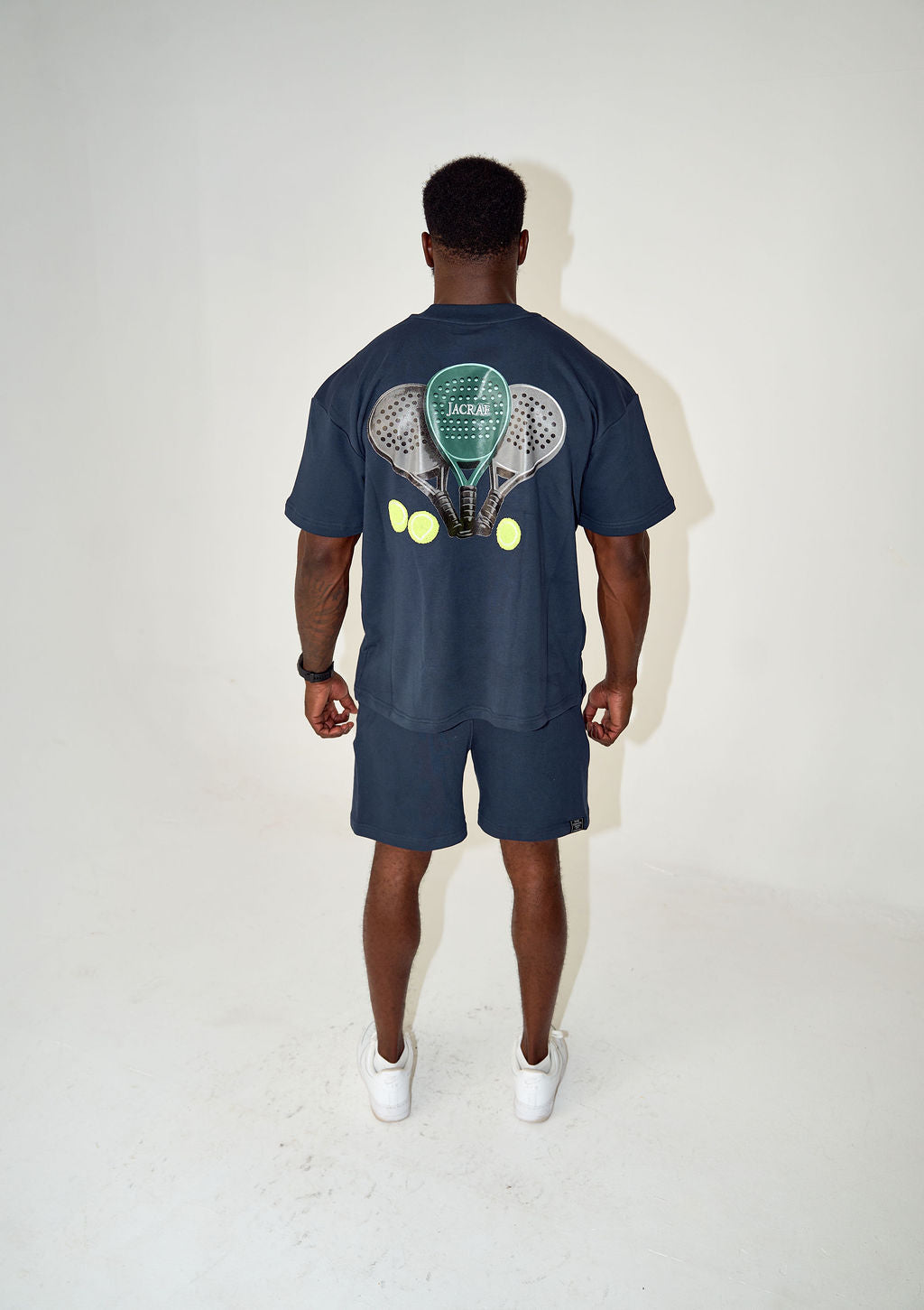 A man viewed from behind wearing a navy blue t-shirt and matching shorts set. The back of the t-shirt features a large graphic print of two tennis rackets with tennis balls.