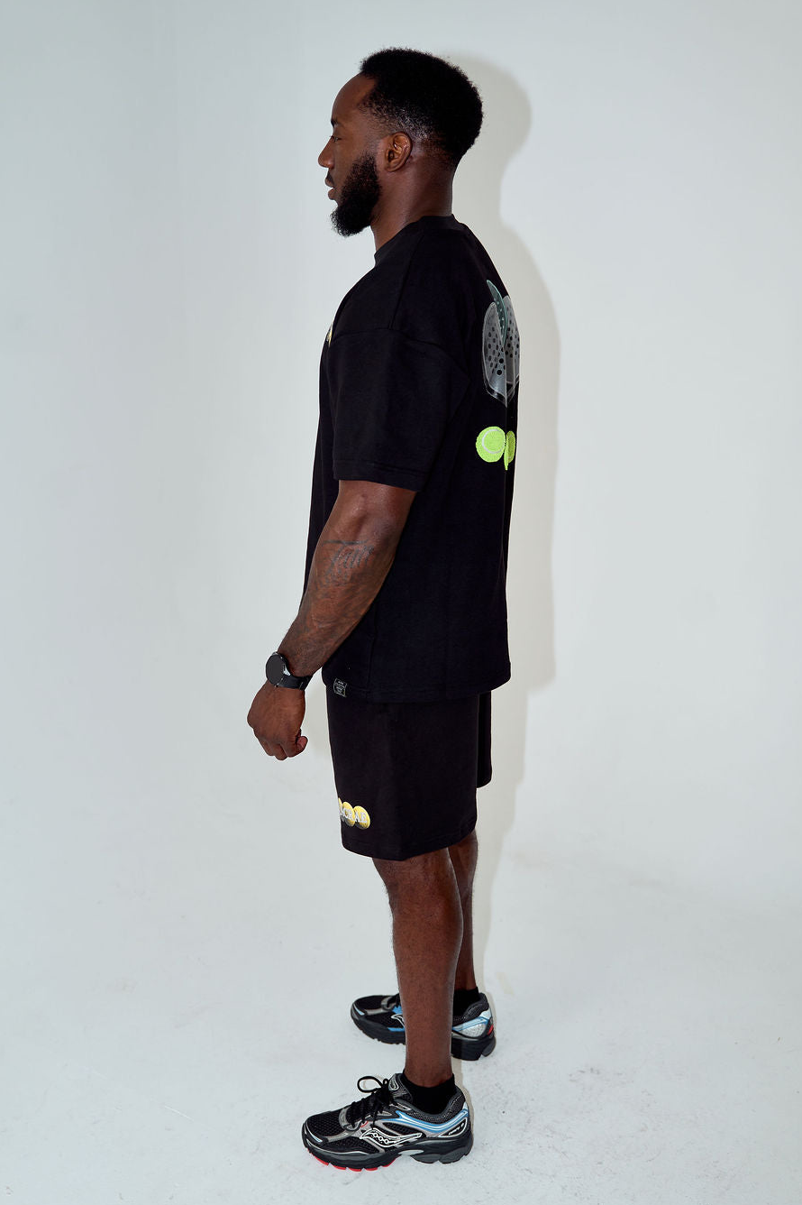 A man shown in profile wearing a black t-shirt and black shorts with small neon yellow-green graphic accents, standing against a plain white studio background.