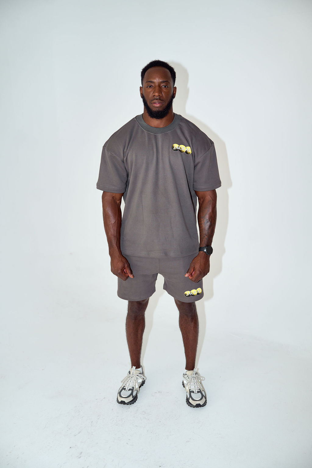 A man stands against a plain white studio background wearing a matching dark gray short-sleeve t-shirt and shorts set with small yellow graphic accents, paired with white and black sneakers.