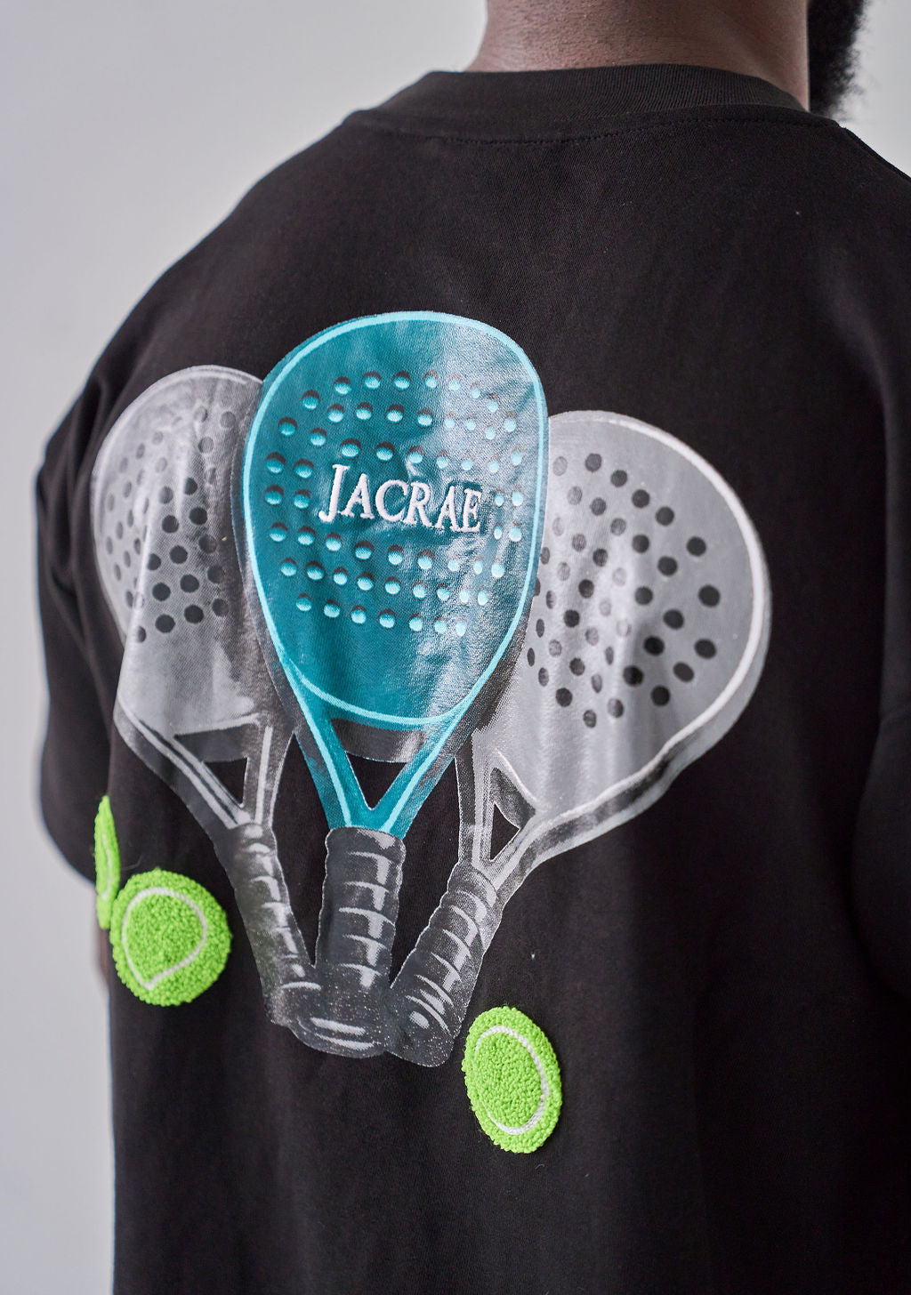 A person wearing a black t-shirt, viewed from the back. The shirt features a large graphic print of three pickleball paddles and two tennis balls, with the word 'JACRAE' printed on the central teal paddle.
