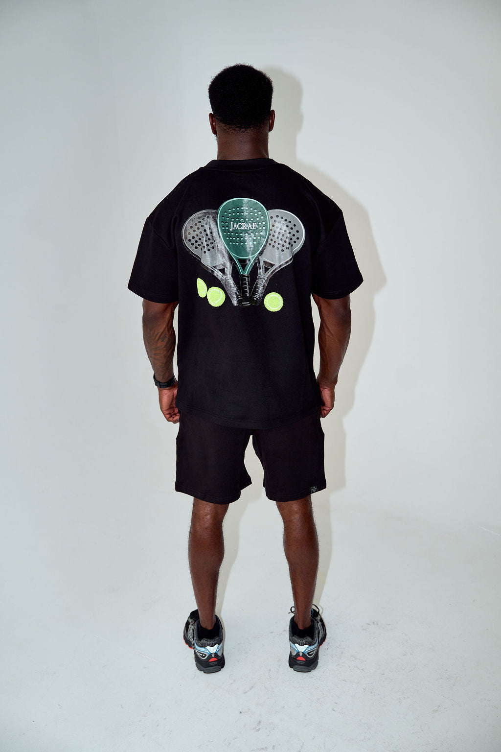 A man viewed from behind wearing a black t-shirt and black shorts, featuring a large graphic print on the back of two tennis rackets and tennis balls, set against a plain white studio background.