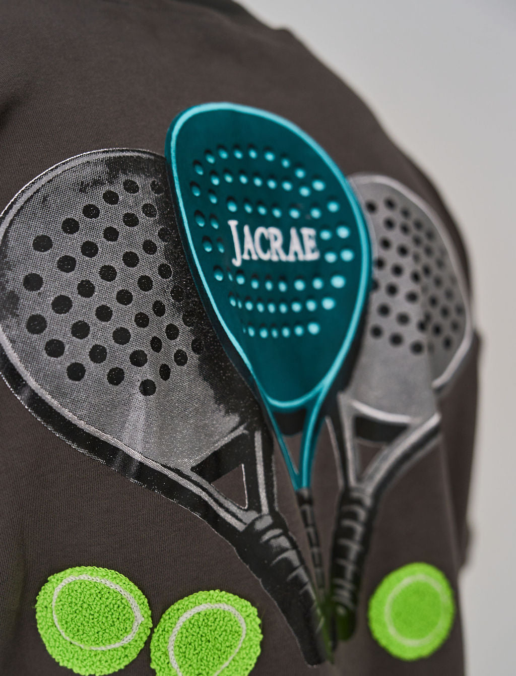 A close-up of a dark brown garment featuring a graphic print of three padel rackets and tennis balls, with a bright teal patch displaying the brand name 'JACRAE' in the center.