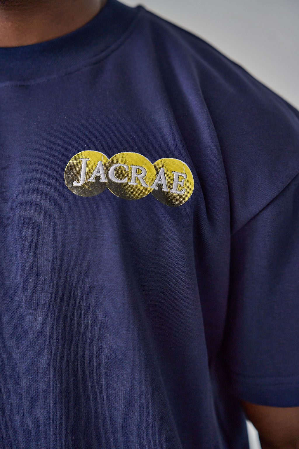 Close-up of a person wearing a navy blue t-shirt with a gold and silver graphic on the chest that reads 'JACRAE'.