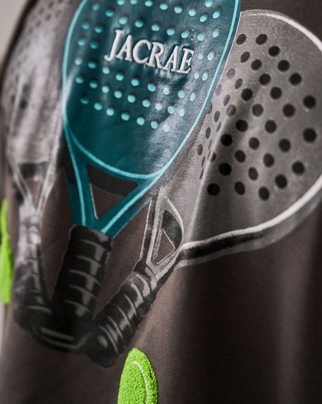 Close-up of a dark gray t-shirt featuring a graphic print of two padel rackets, one teal and one gray, with the brand name 'JACRAE' printed on the teal racket head.