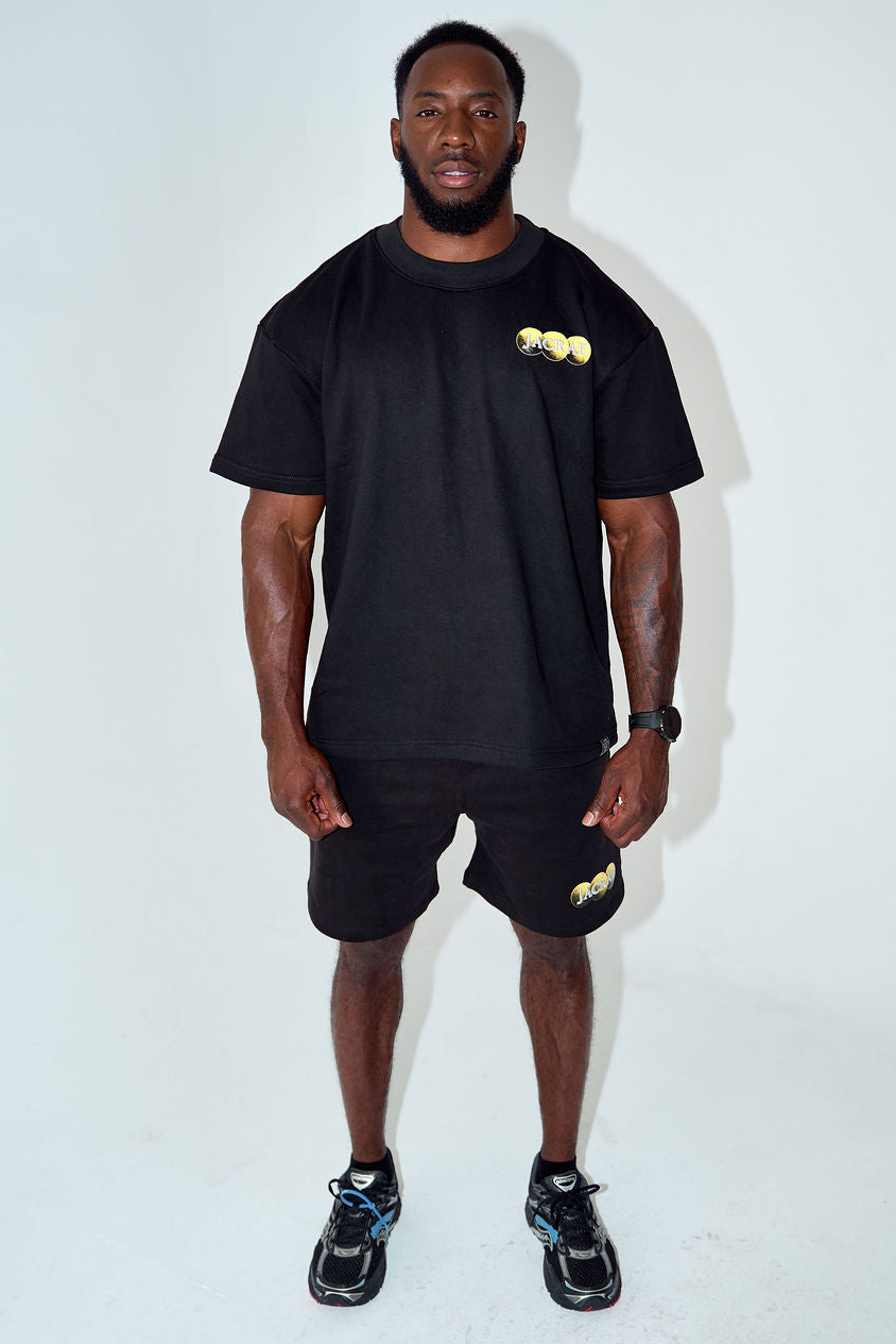 A man modeling a black short-sleeve t-shirt and matching black shorts with a small gold graphic on the chest and thigh, paired with black sneakers, against a plain white studio background.