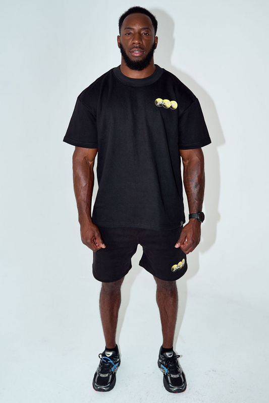 A man modeling a black short-sleeve t-shirt and matching black shorts with a small gold graphic on the chest and thigh, paired with black sneakers, against a plain white studio background.