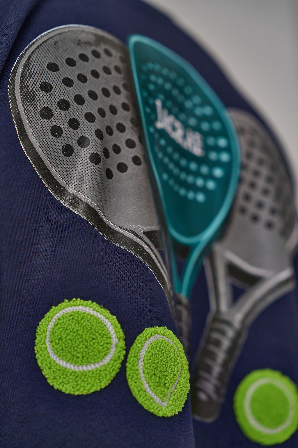 A close-up of a navy blue garment featuring a large graphic print of padel rackets and tennis balls, including two green and white textured tennis balls near the bottom left.